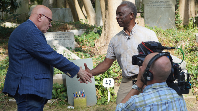 Public TV Series: World's Greatest Cemeteries. Episode 102. London, UK. Highgate Cemetery. (left to right) Docent Peter Mills, Cameraman Richard Reed and Host/Producer Roberto Mighty at the grave of writer Douglas Adams, author of "The Hitchhiker's Guide to The Galaxy". ©2019 Roberto Mighty. Photo by Kate Taylor Mighty